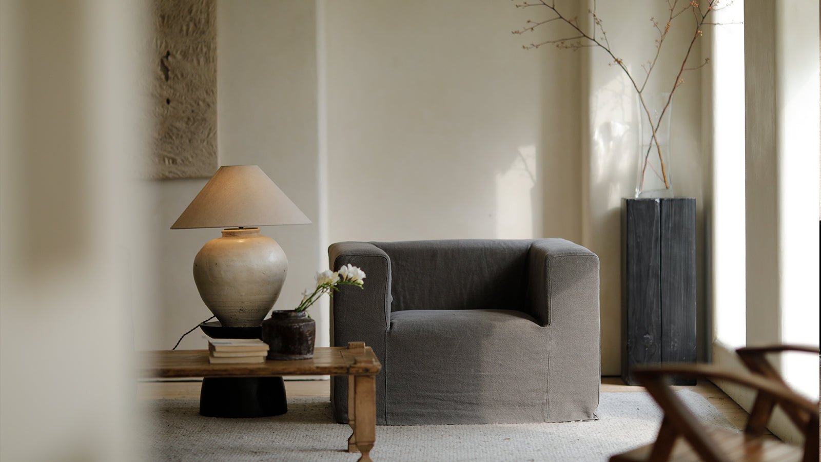 A modern grey armchair with a slipcover, positioned next to a wooden side table with a large, stylish ceramic lamp