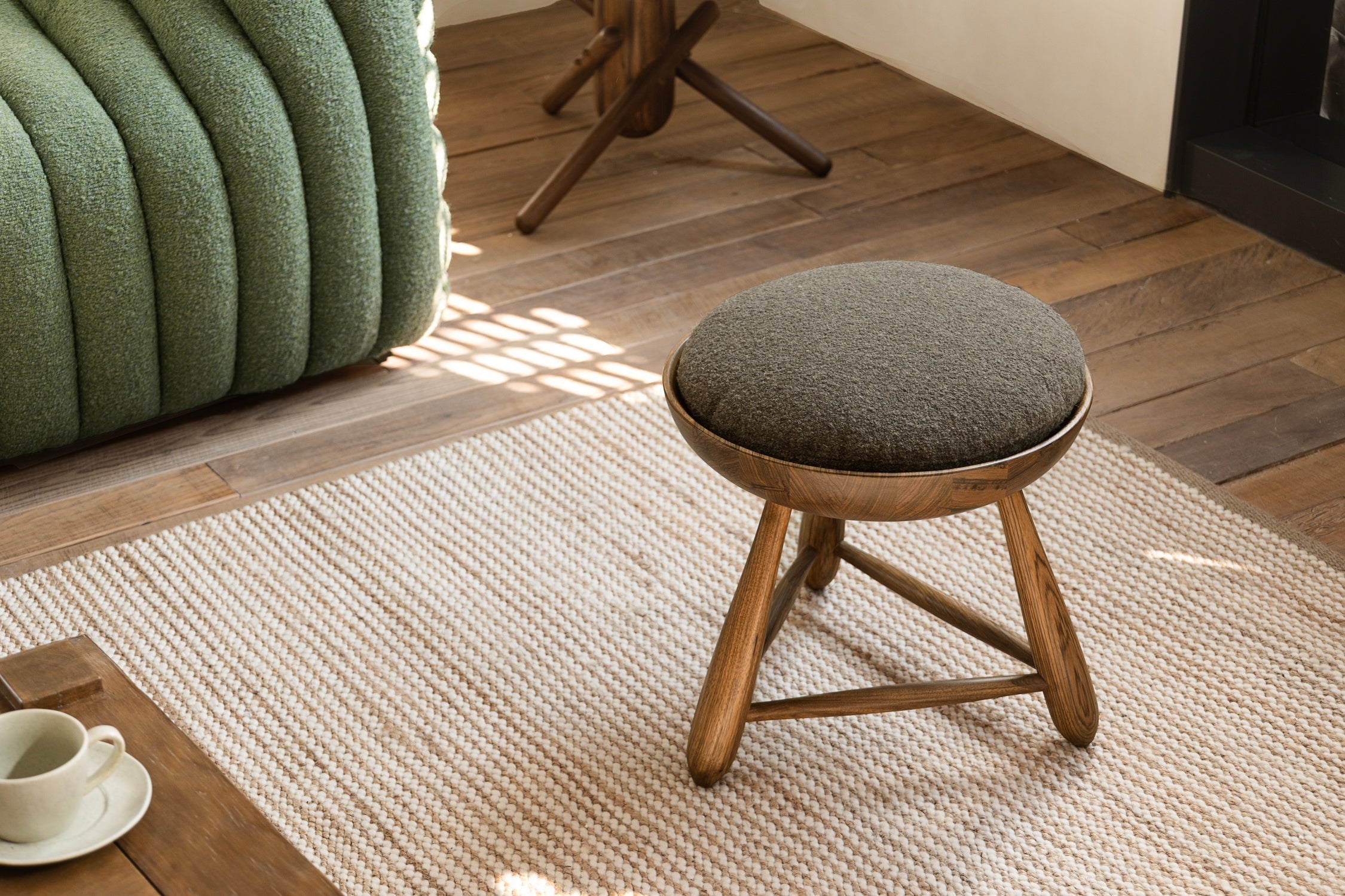 A round cushioned stool resting on a woven rug beside a green textured sofa in a sunlit space.