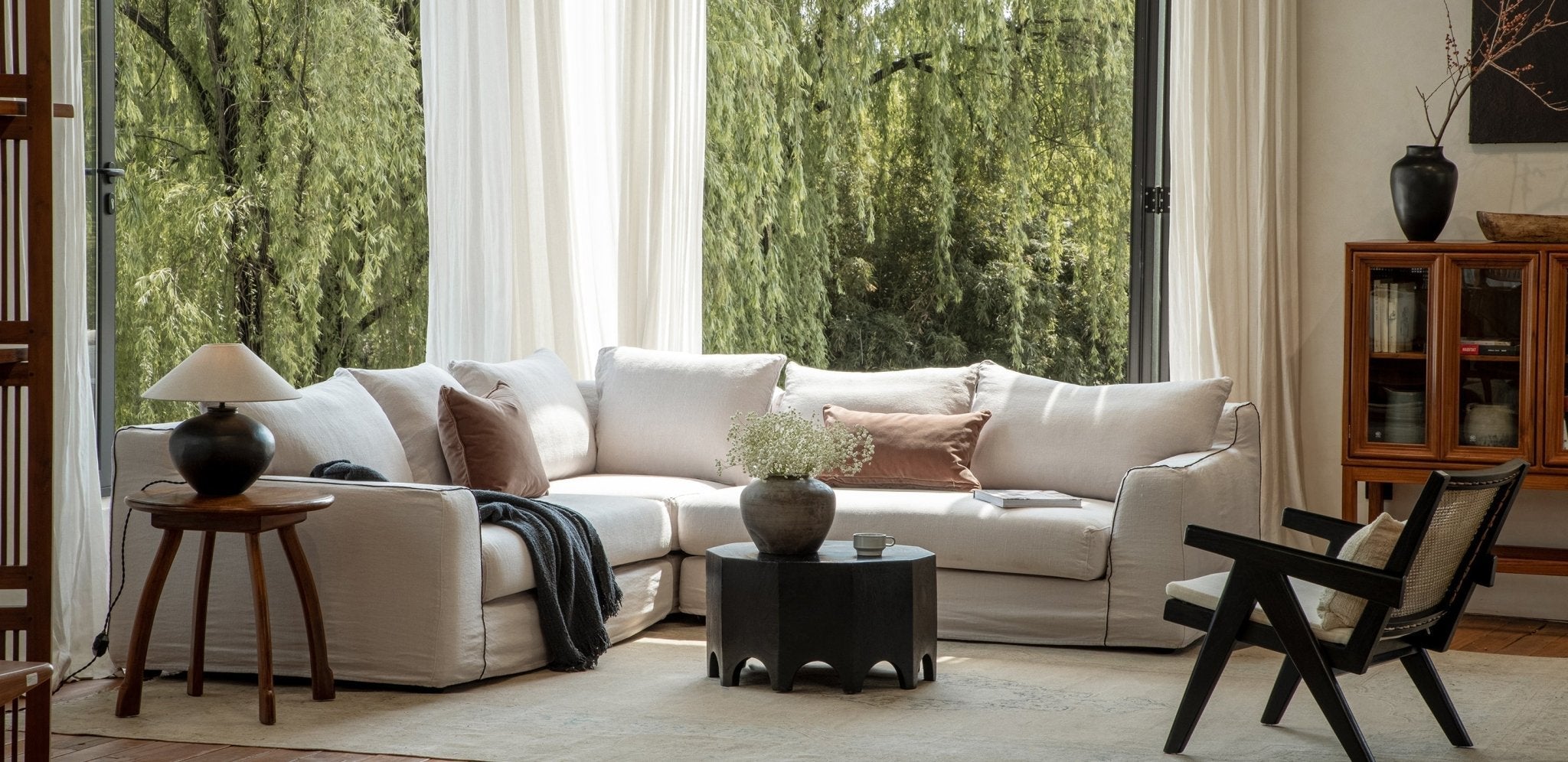 A bright living room featuring a white sectional sofa with cushions, surrounded by wooden furniture and large windows overlooking greenery.