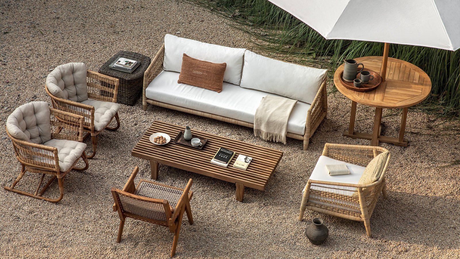 An outdoor lounge set on gravel, featuring a sofa, rattan armchairs, a coffee table, and a large white umbrella