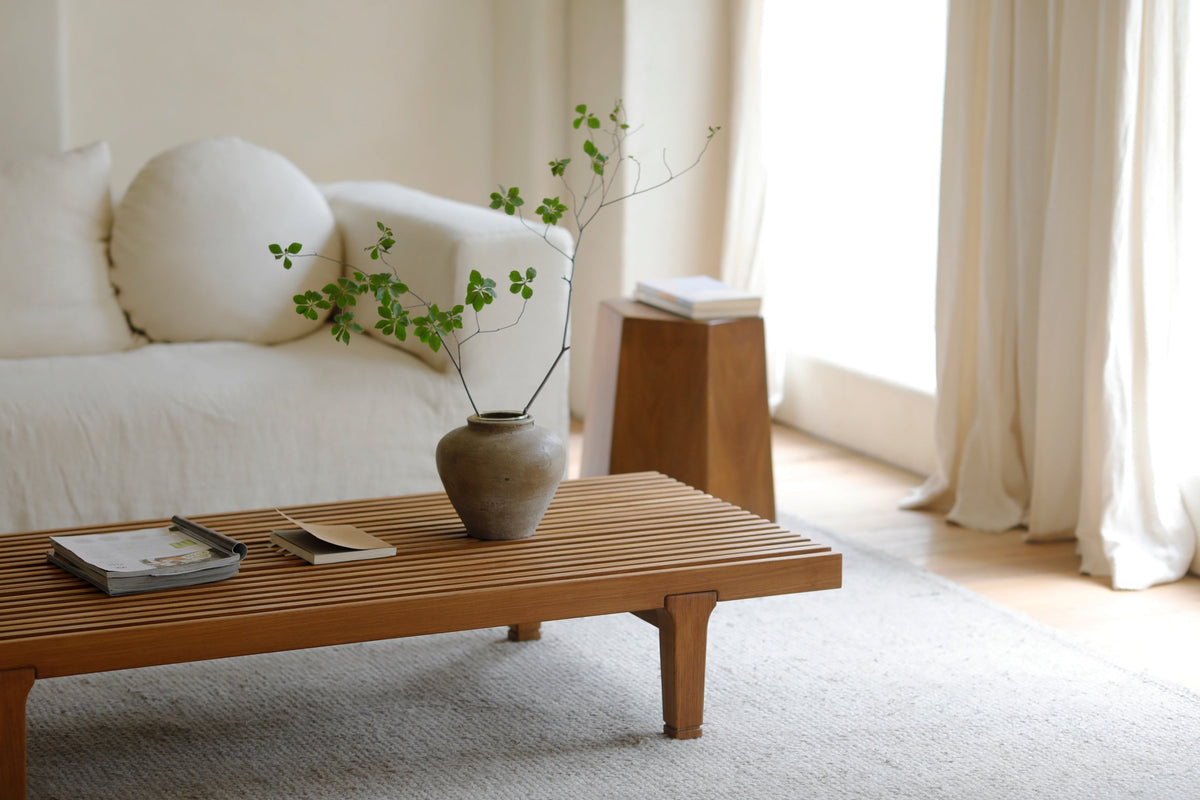 A low slatted wooden coffee table displaying a leafy vase and books in a bright, minimalist living room.