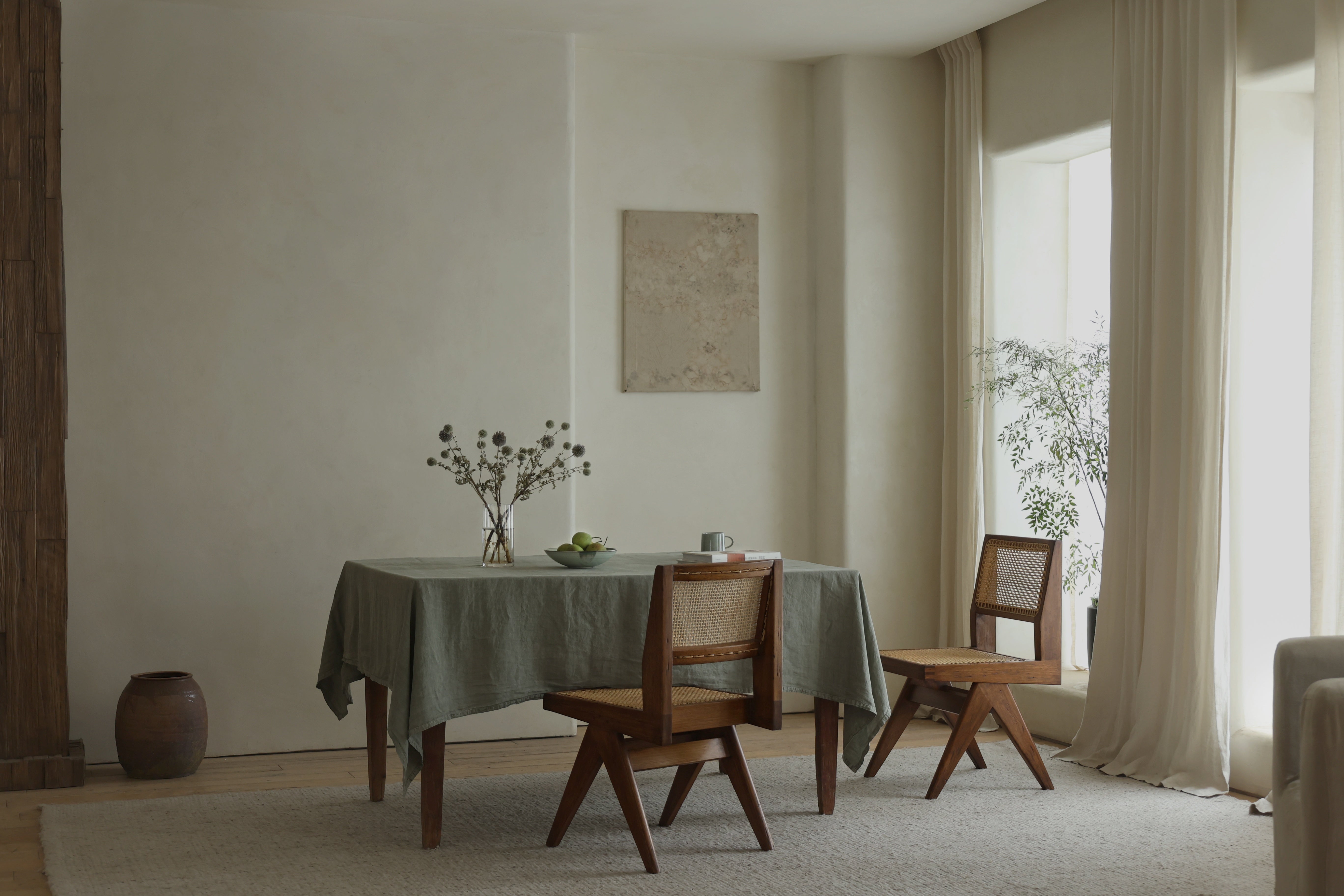 Two wooden dining chairs with rattan backrests are placed beside a table draped with a soft, sage-green tablecloth in a serene dining space.