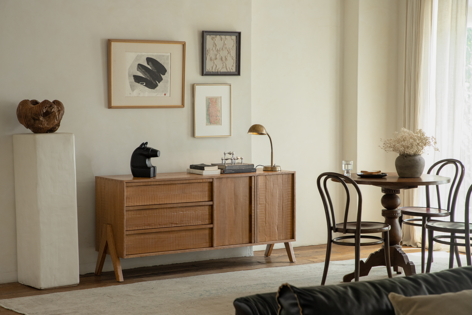 A cozy dining area with a round wooden table, bentwood chairs, and a wooden sideboard decorated with art and a lamp.
