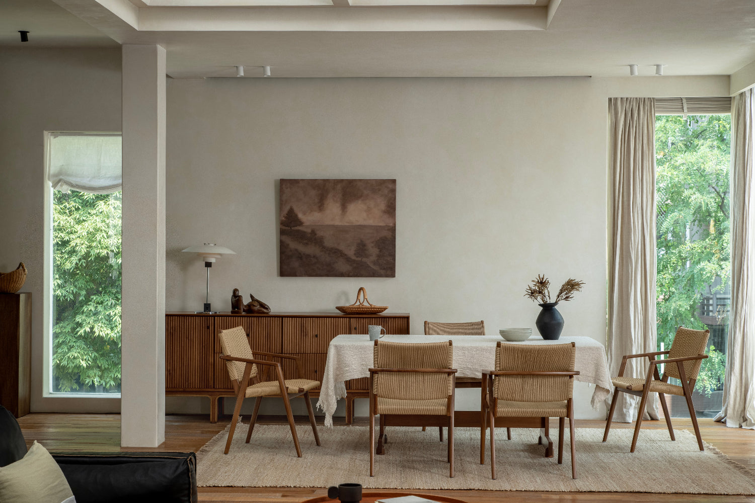 A dining table set with chairs in a bright room with large windows and a sideboard against the wall.