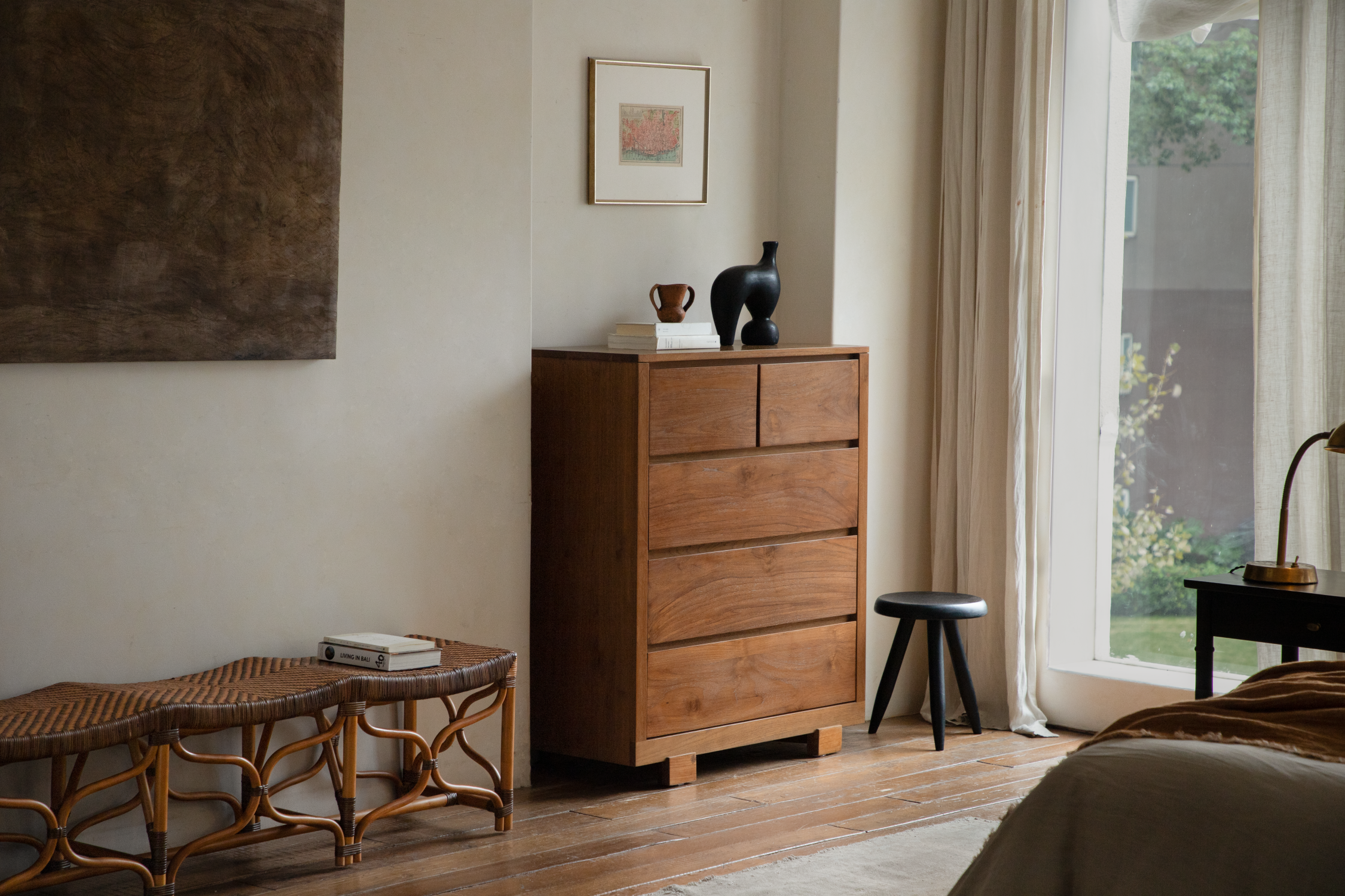 A minimalist bedroom interior featuring a wooden chest of drawers, a woven bench, and neutral decor in a warm, earthy color palette.