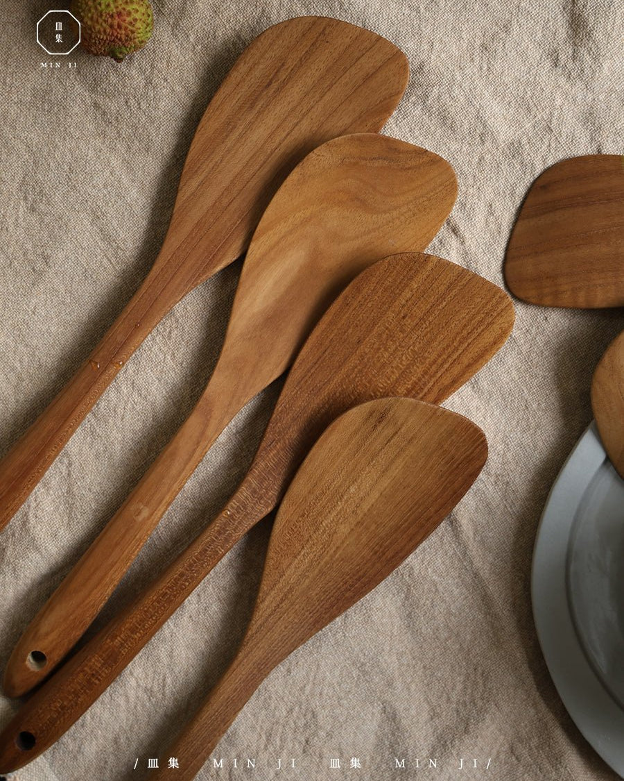 Natural teak wood fried spatulas (Variants A & B) with smooth, curved heads on linen fabric.
