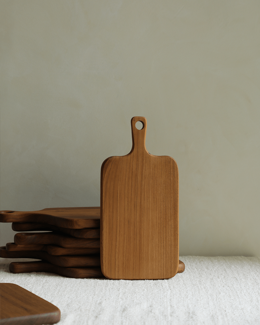 Handcrafted teak cutting board with handle, stack of boards behind.