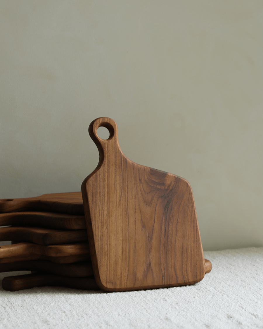 Handcrafted teak cutting board with looped handle, stacked on textured linen.