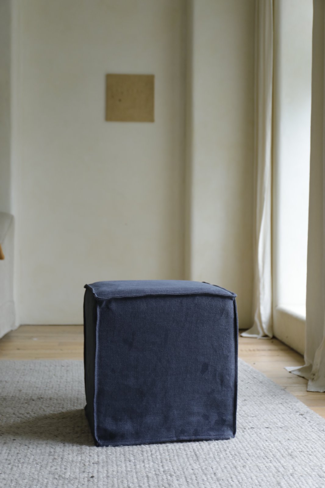 A Cube Linen Ottoman by ZM Home in dark blue sits on a light textured rug in a minimalist room with pale walls and natural window light.