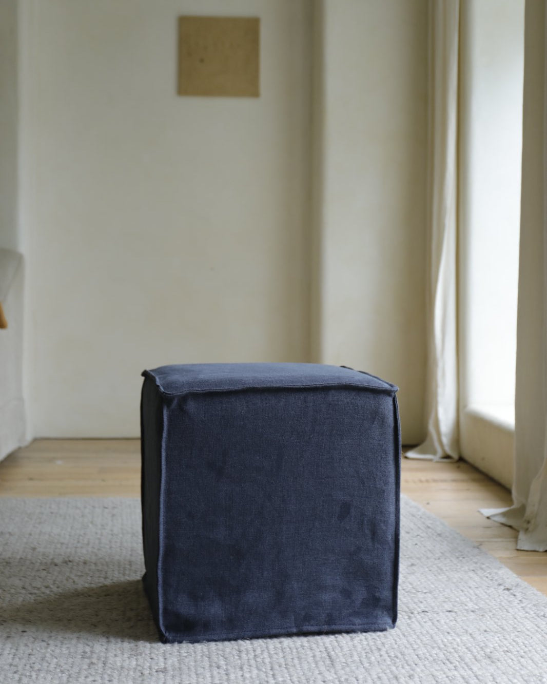 A Cube Linen Ottoman by ZM Home in dark blue sits on a light textured rug in a minimalist room with pale walls and natural window light.