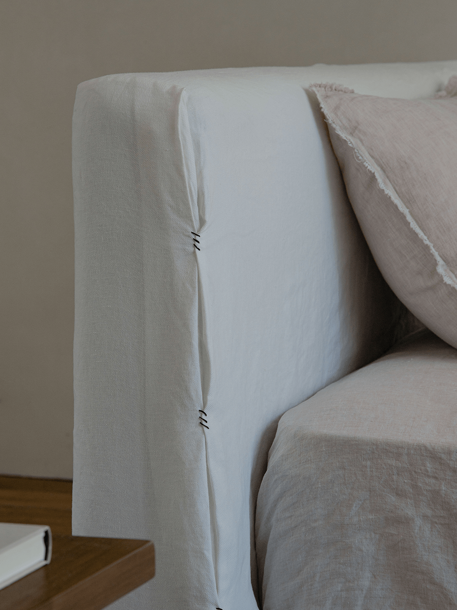 Freya Linen Bed headboard detail: White linen upholstery, stitching, and a soft pink pillow for peaceful sleep.