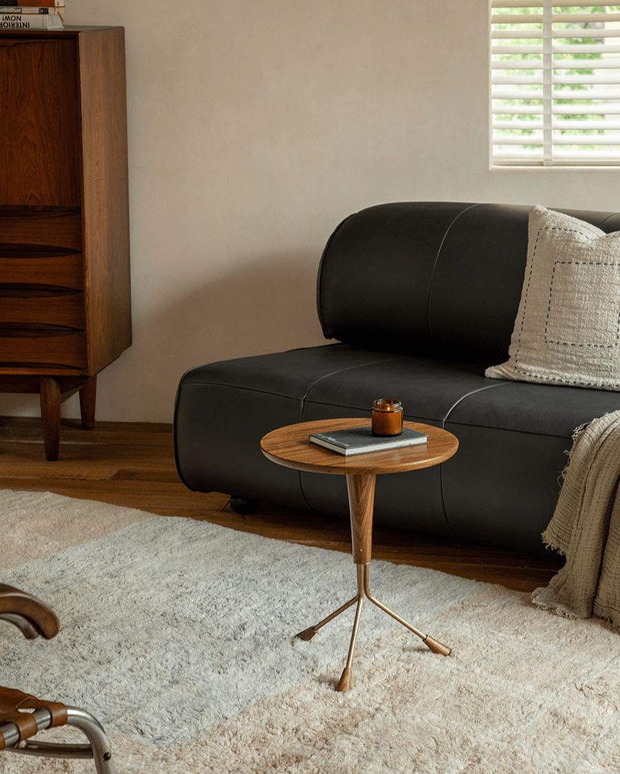 Larsson Side Table with ebony wood top and brass tripod base next to a black leather sofa