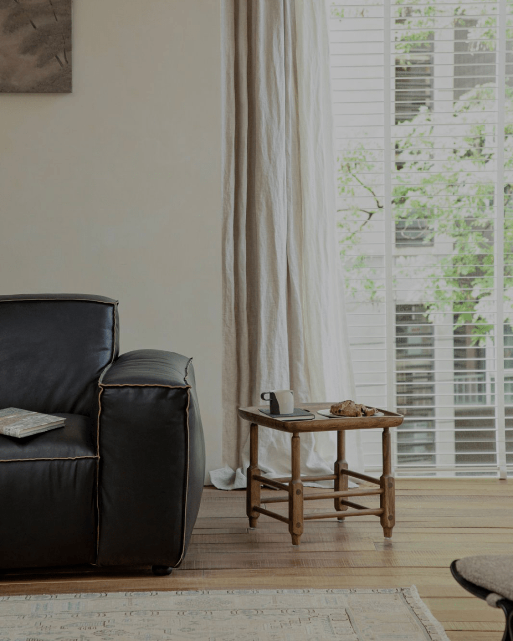 Modern living room with black leather armchair, wooden side table, and Magrini stool, natural light.