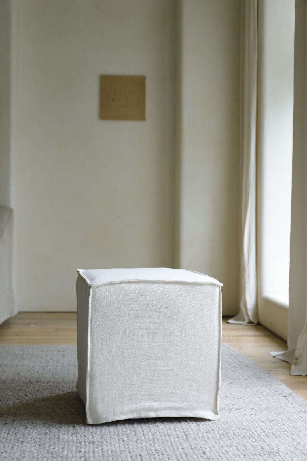 The ZM Home Cube Linen Ottoman rests on a textured gray rug in a minimalist room with light walls and a large window.