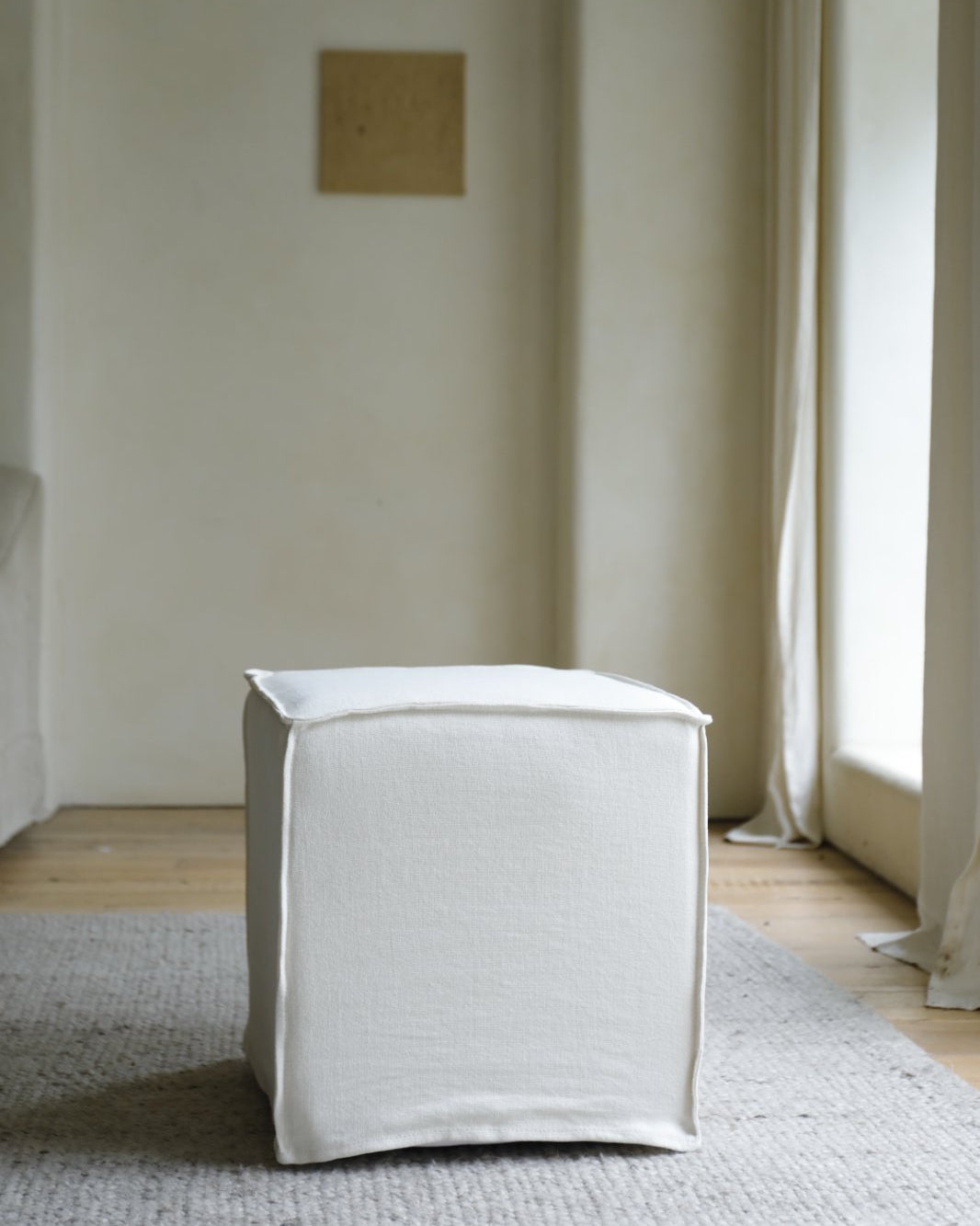 The ZM Home Cube Linen Ottoman rests on a textured gray rug in a minimalist room with light walls and a large window.