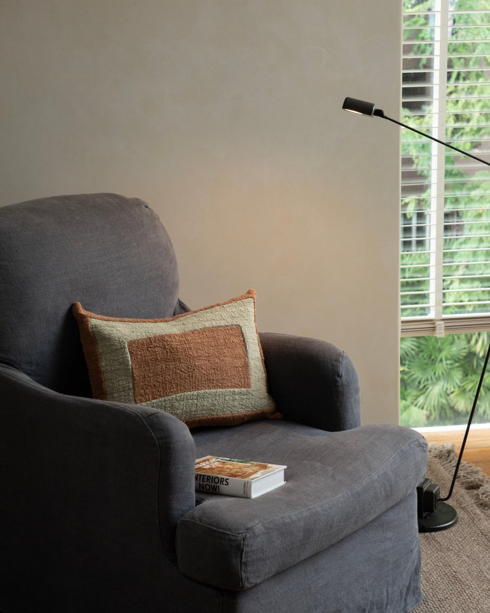 Handwoven Sylia pillow in rust and sage on a dark grey armchair, with a book titled 'Interiors Now!' and a floor lamp.