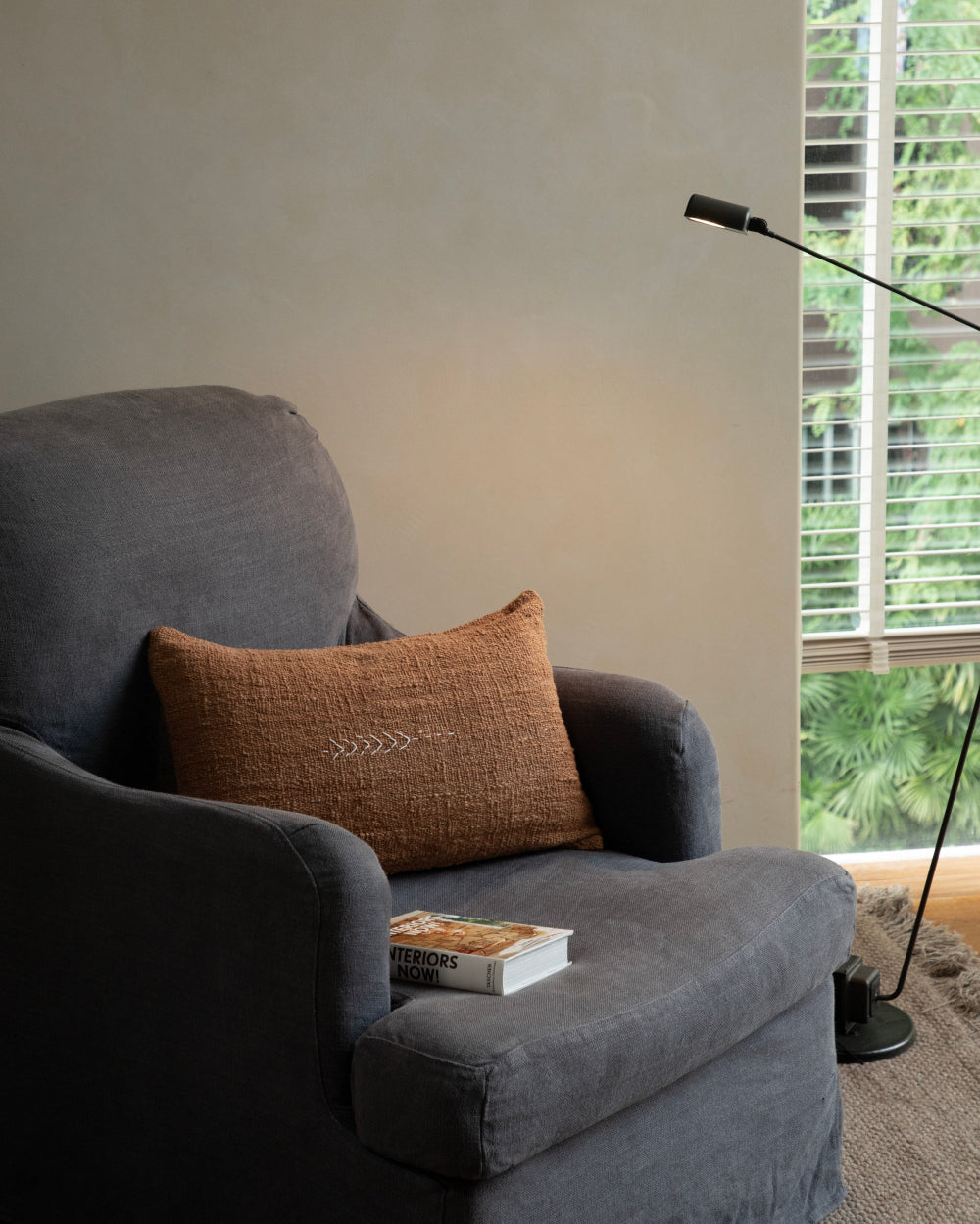 Sylia woven lumbar pillow in earthy rust with white stitching, on charcoal armchair. Book nearby.