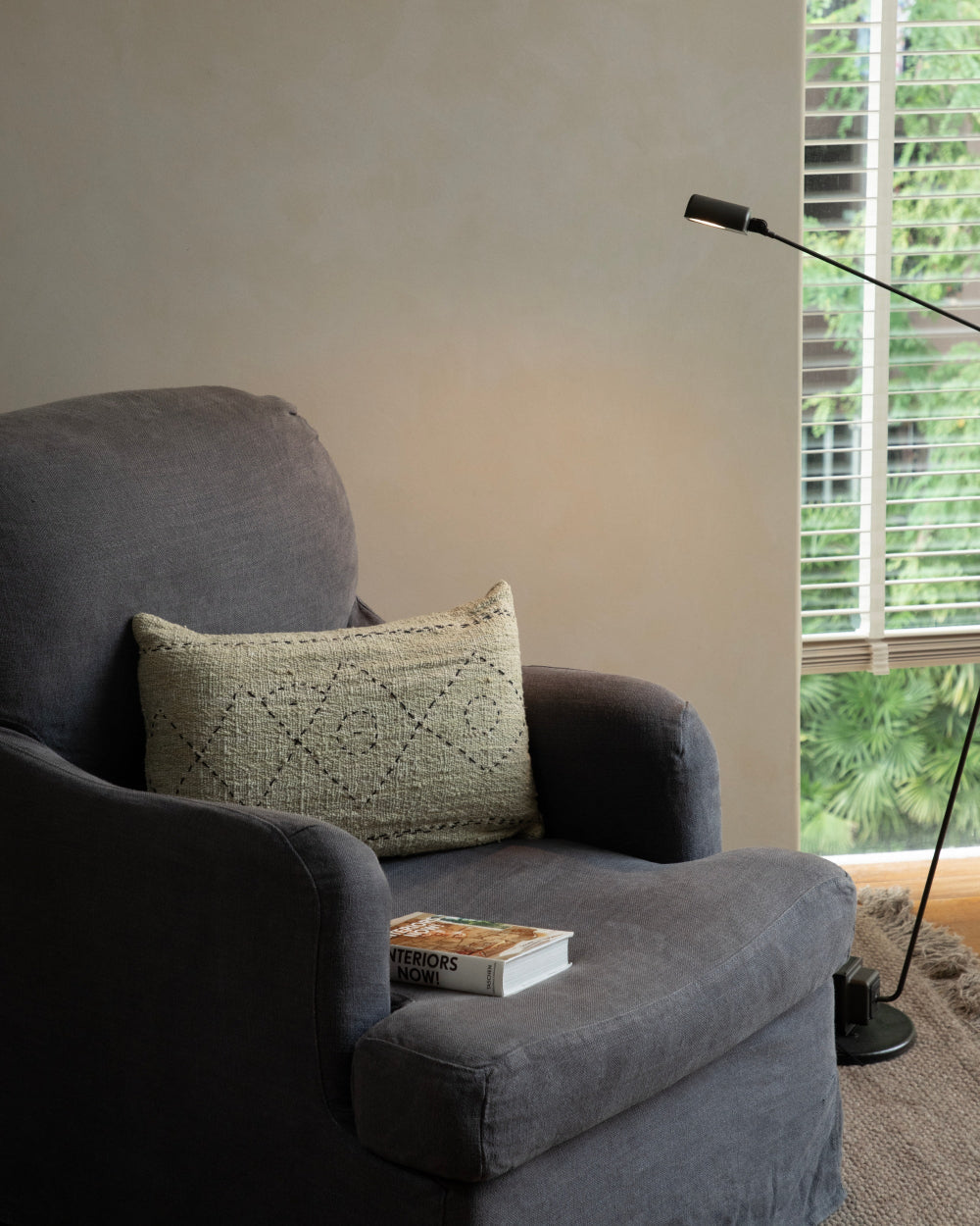 Soft, handwoven Sylia Pillow with diamond pattern on a dark gray armchair, near a book and modern lamp.