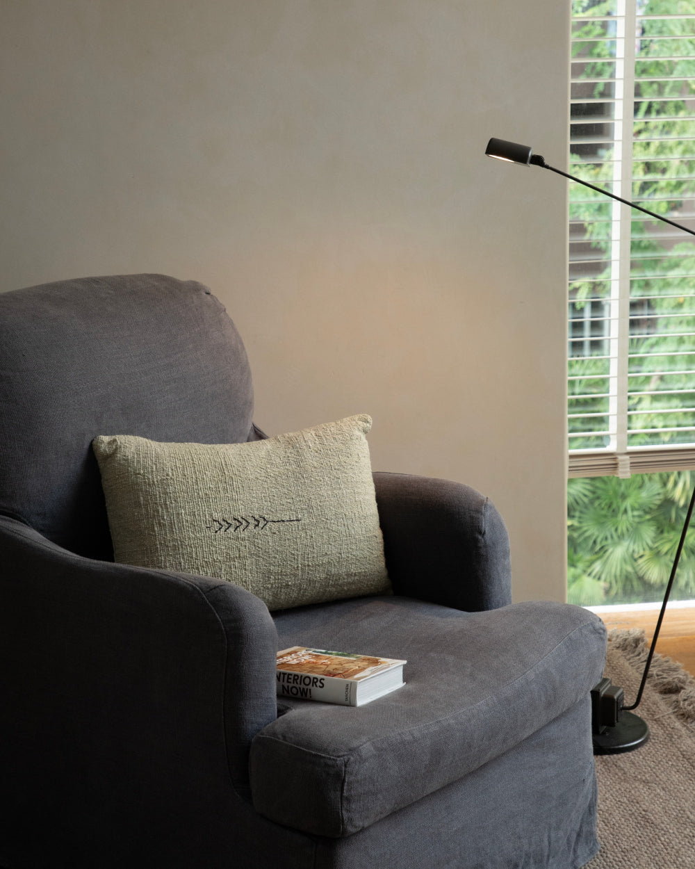 Handwoven Sylia Pillow on armchair with subtle natural color and embroidered detail, beside 'Interiors Now!' book.