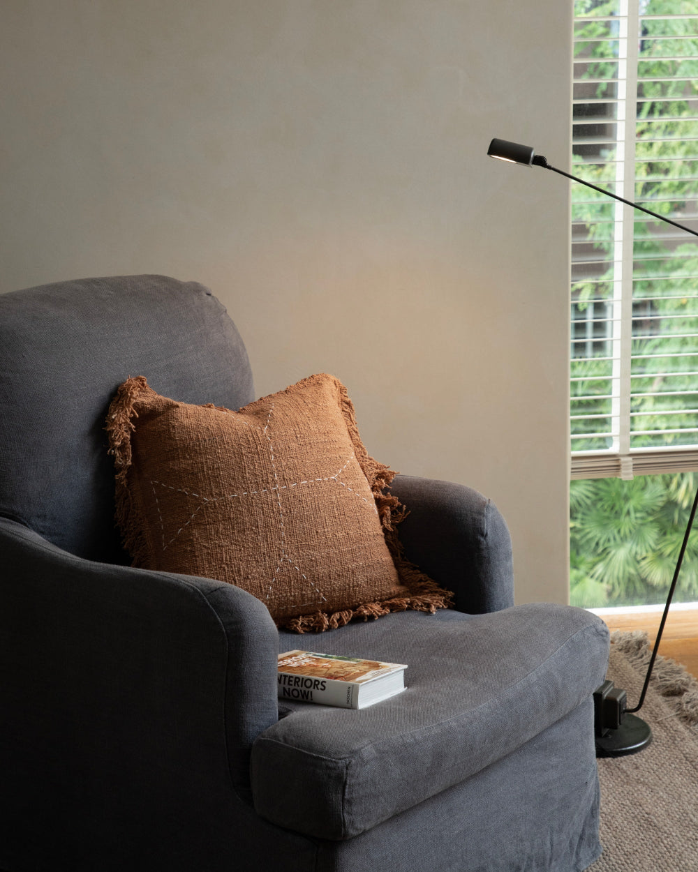Handwoven Sylia Pillow in autumn leaf color on a charcoal armchair with a book.