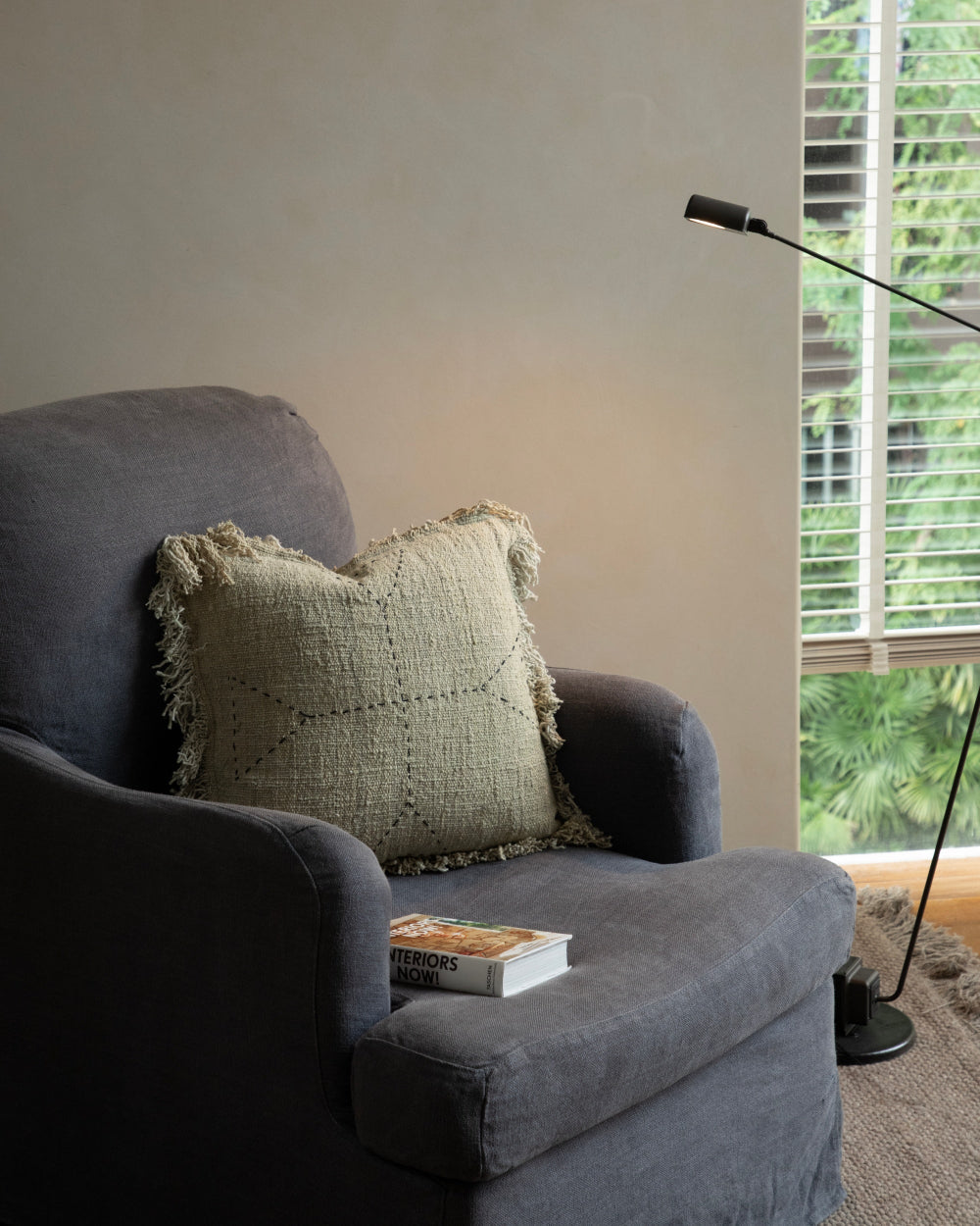 Handwoven Sylia Pillow with fringe on a grey armchair, featuring a subtle stitched design in calming neutral tones.