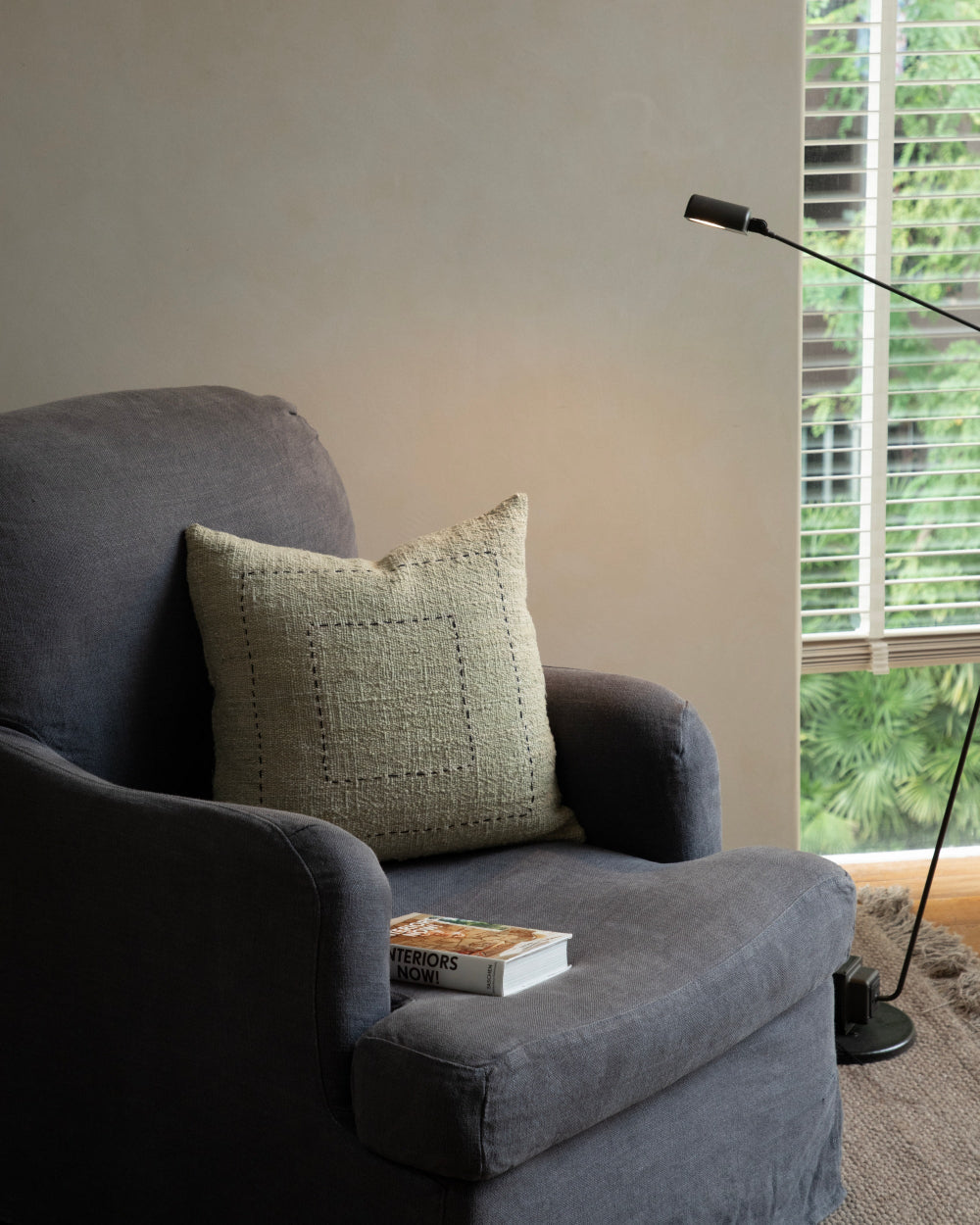 Handwoven Sylia Pillow in a spring grass shade on a charcoal armchair, with "Interiors Now!" book.