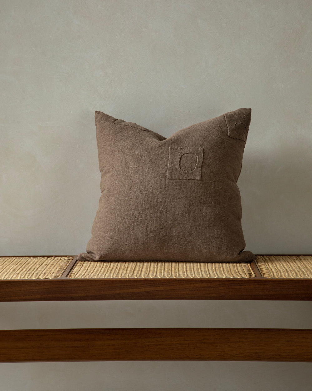 A brown, textured linen pillow with a small stitched square detail sits on a wooden bench with a cane seat.
