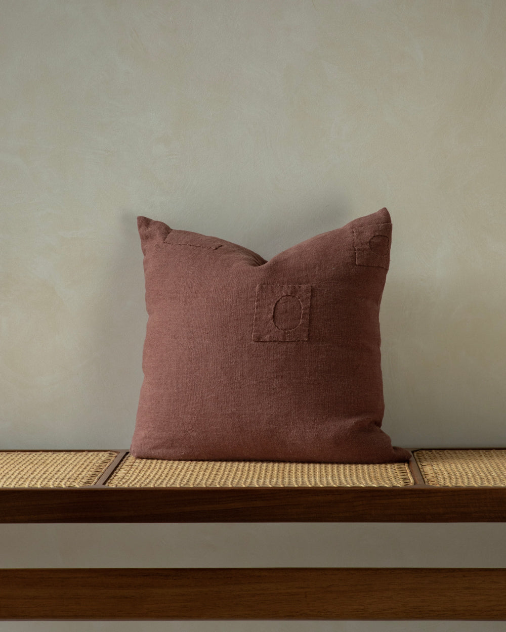 Woven linen pillow in tea brown with stitched square details, displayed on a wooden bench with woven seat.