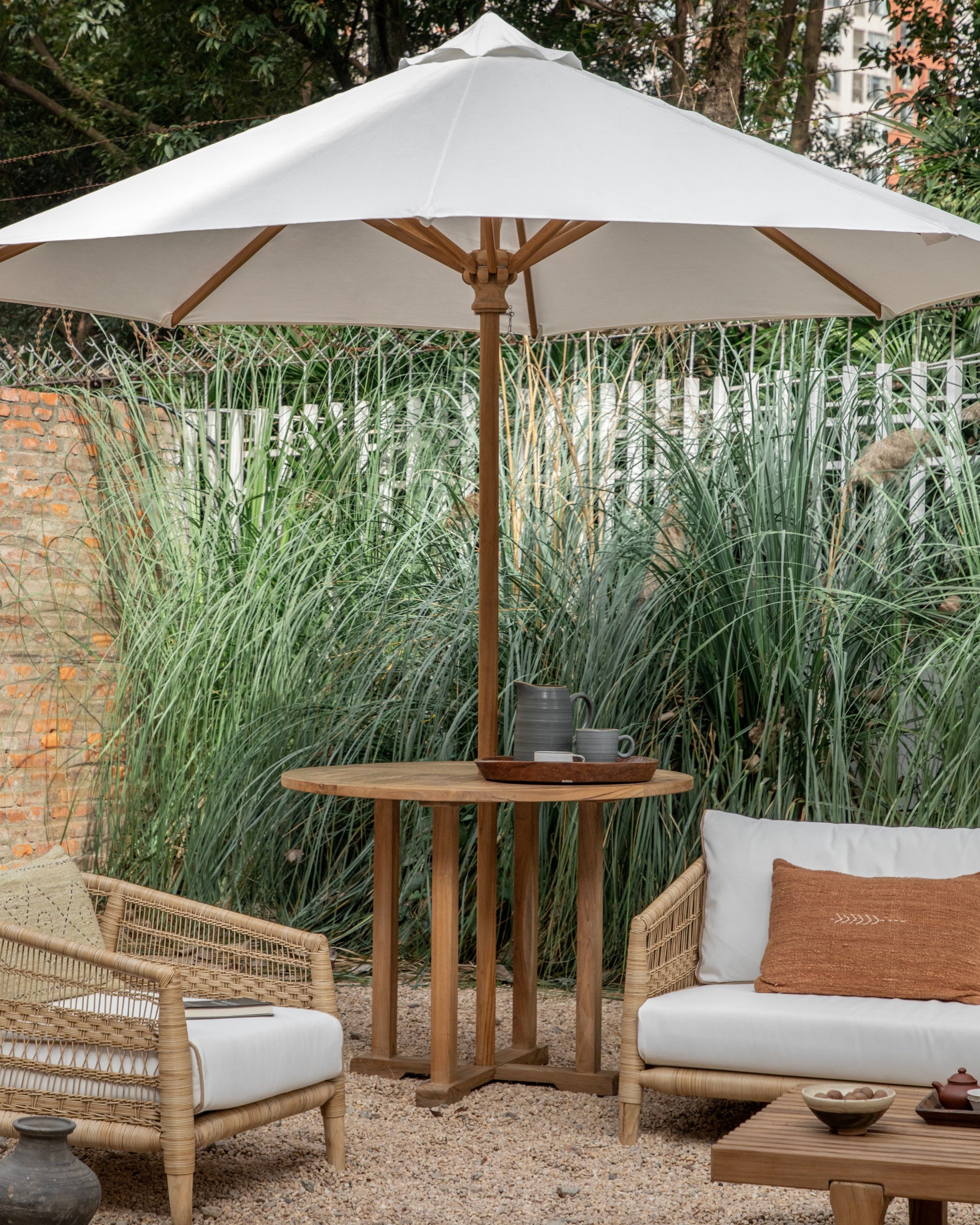 Teak outdoor patio table and white umbrella set with wicker chairs, surrounded by greenery.