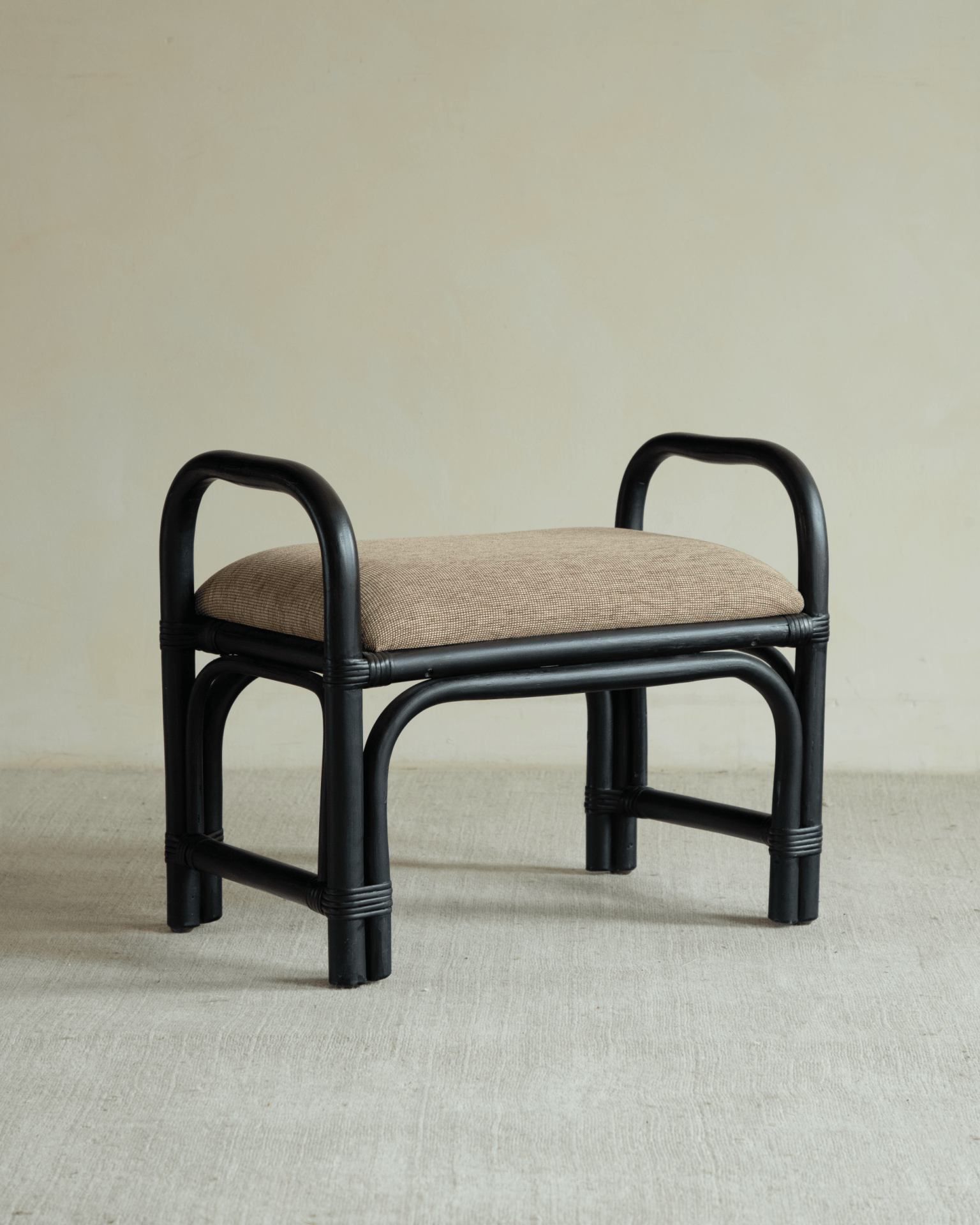 Black rattan stool with beige linen-cotton seat and curved handles.