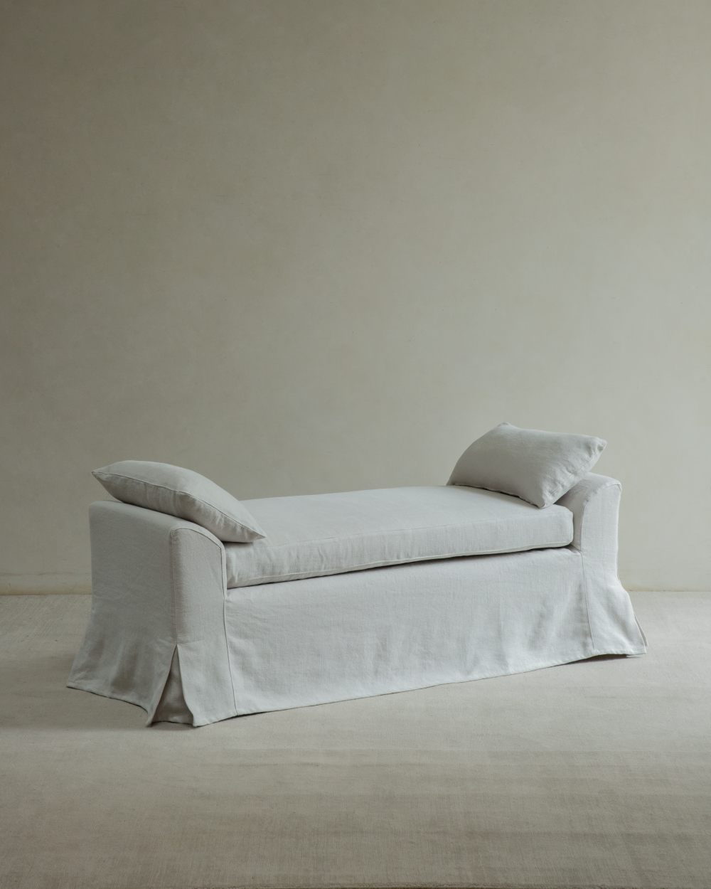 Alina Bench in Linen Gray with slipcover, two pillows, wooden legs, and skirted base on a rug.