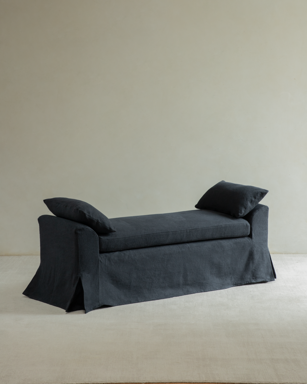 Alina Bench upholstered in dark yarn-dyed linen with a flowing skirted base and two accent pillows.