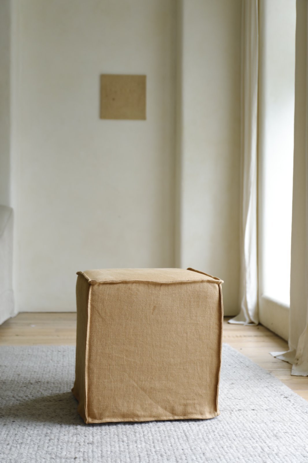 The ZM Home Cube Linen Ottoman rests on a textured rug in a minimalist room with light walls and large windows.