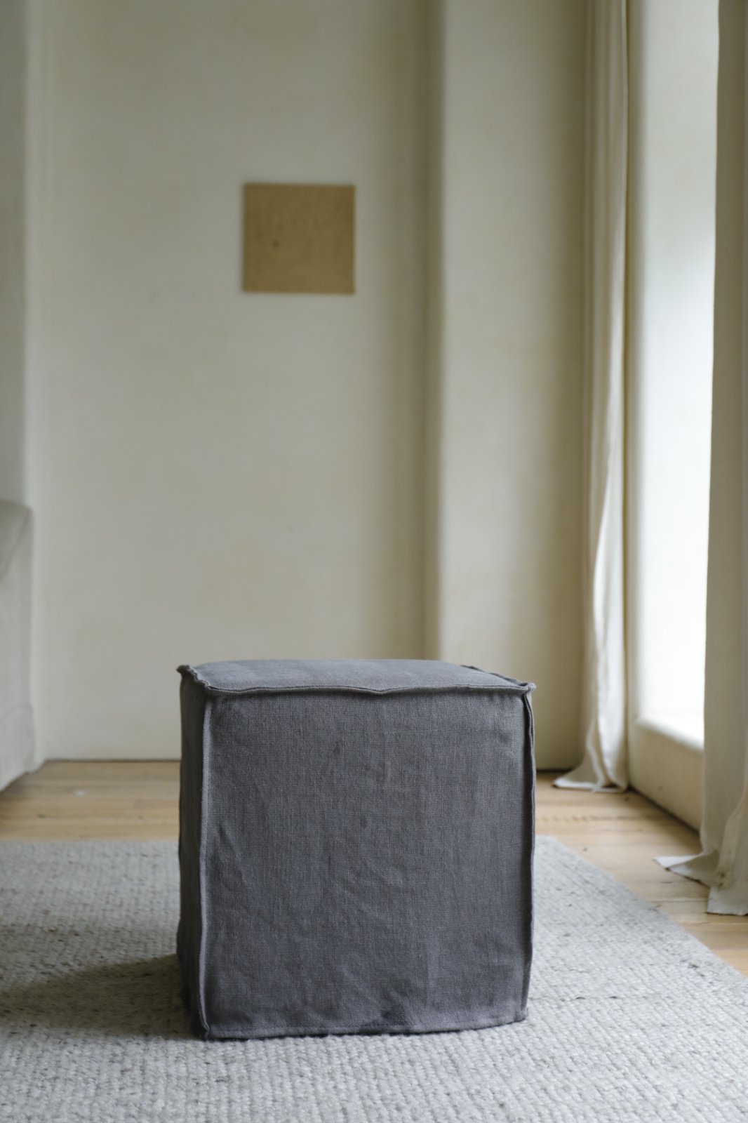 The ZM Home Cube Linen Ottoman in gray rests on a light textured rug in a minimalist room with cream walls, tall windows, and a small square artwork on the wall.