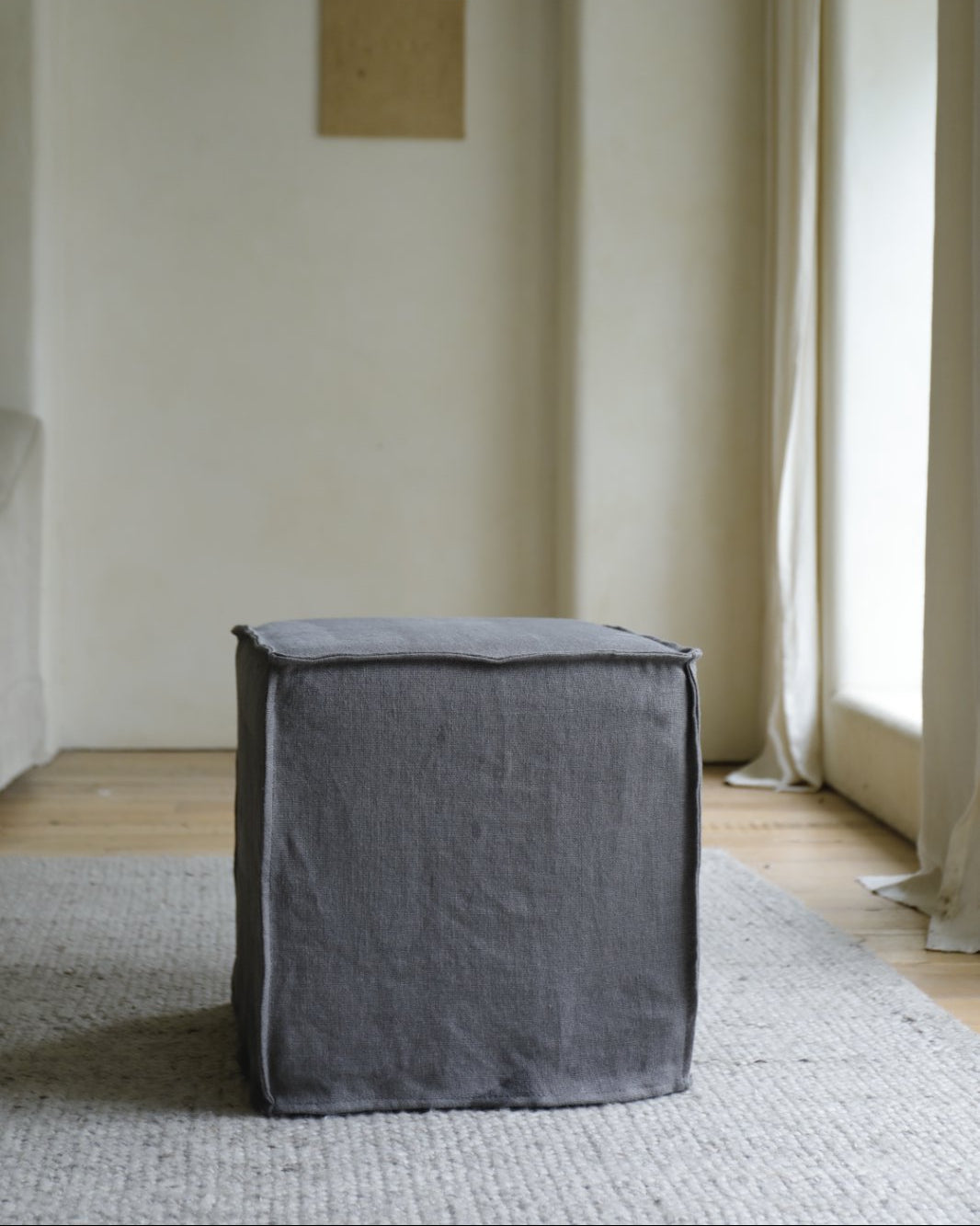 The ZM Home Cube Linen Ottoman in gray rests on a light textured rug in a minimalist room with cream walls, tall windows, and a small square artwork on the wall.