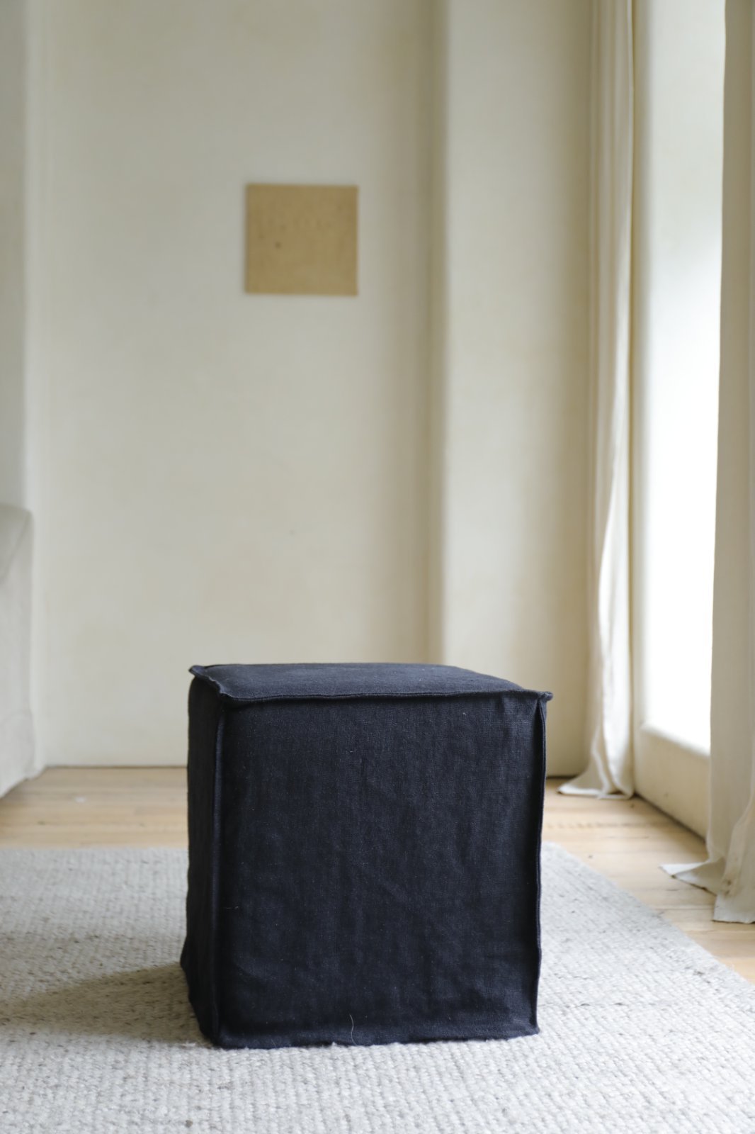 The Cube Linen Ottoman by ZM Home, in black fabric, rests on a light textured rug in a minimalist room with cream walls and large windows.