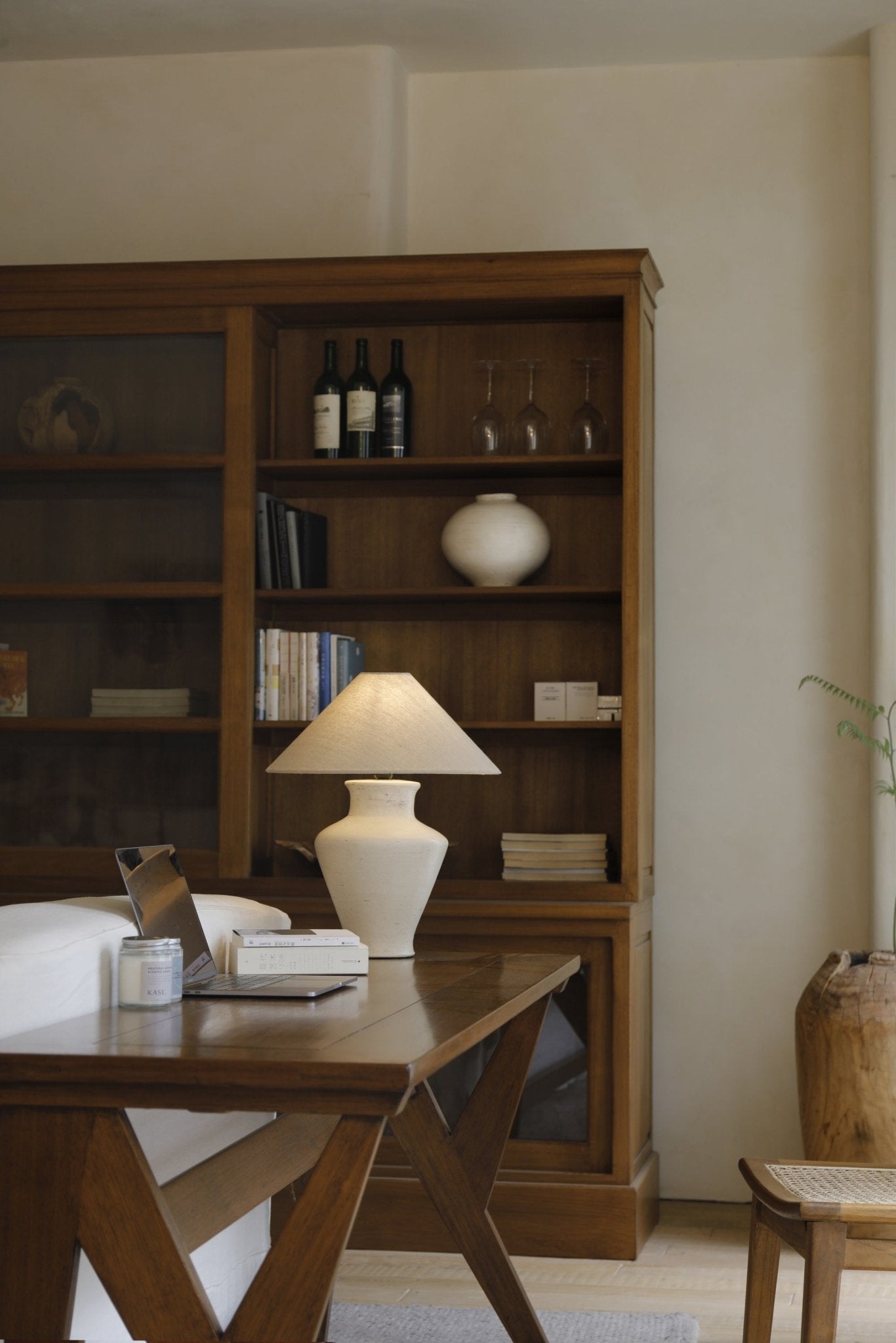Short neck lamp on a wooden table with a clay base and linen shade, next to a laptop, books and wine.