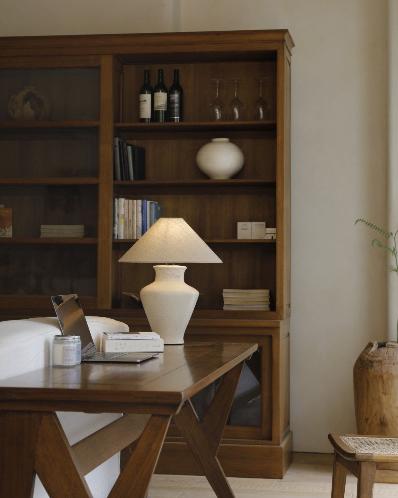 Short neck lamp on a wooden table with a clay base and linen shade, next to a laptop, books and wine.