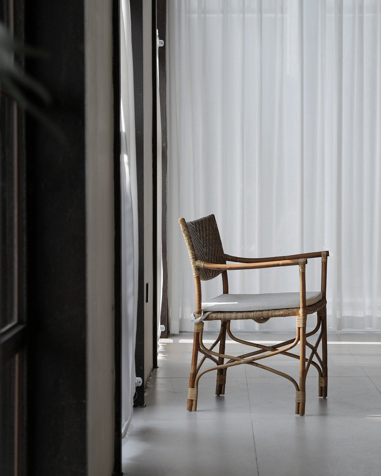 Dingee Chair with woven back, natural wood frame, and cushion, next to sheer white curtains in a bright room