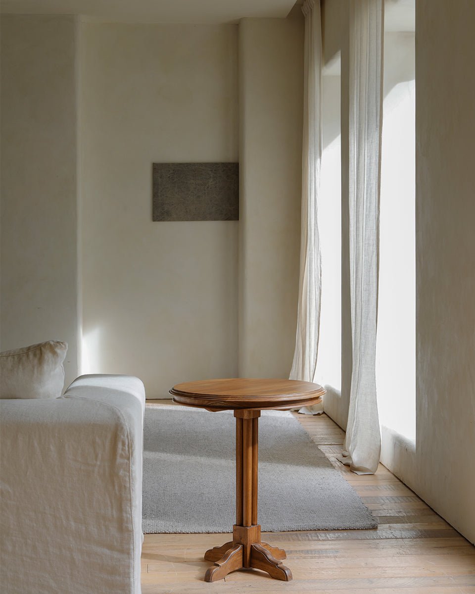 Round Indonesian teak side table, detailed pedestal base, geometric design, next to white sofa in minimalist interior.