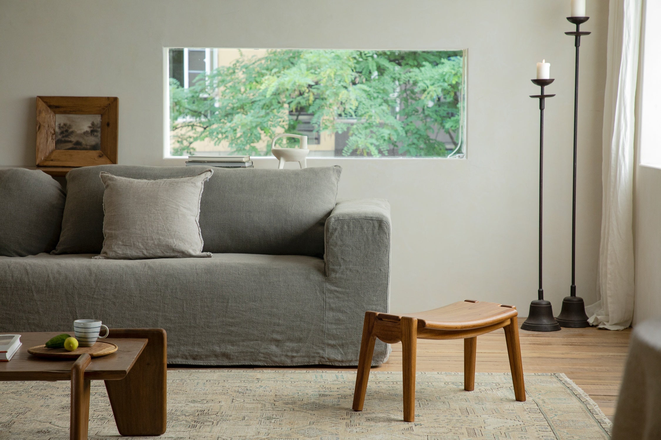 A small wooden stool stands on a rug in front of a grey sofa, positioned near two tall candle holders in a bright, minimalist living room.