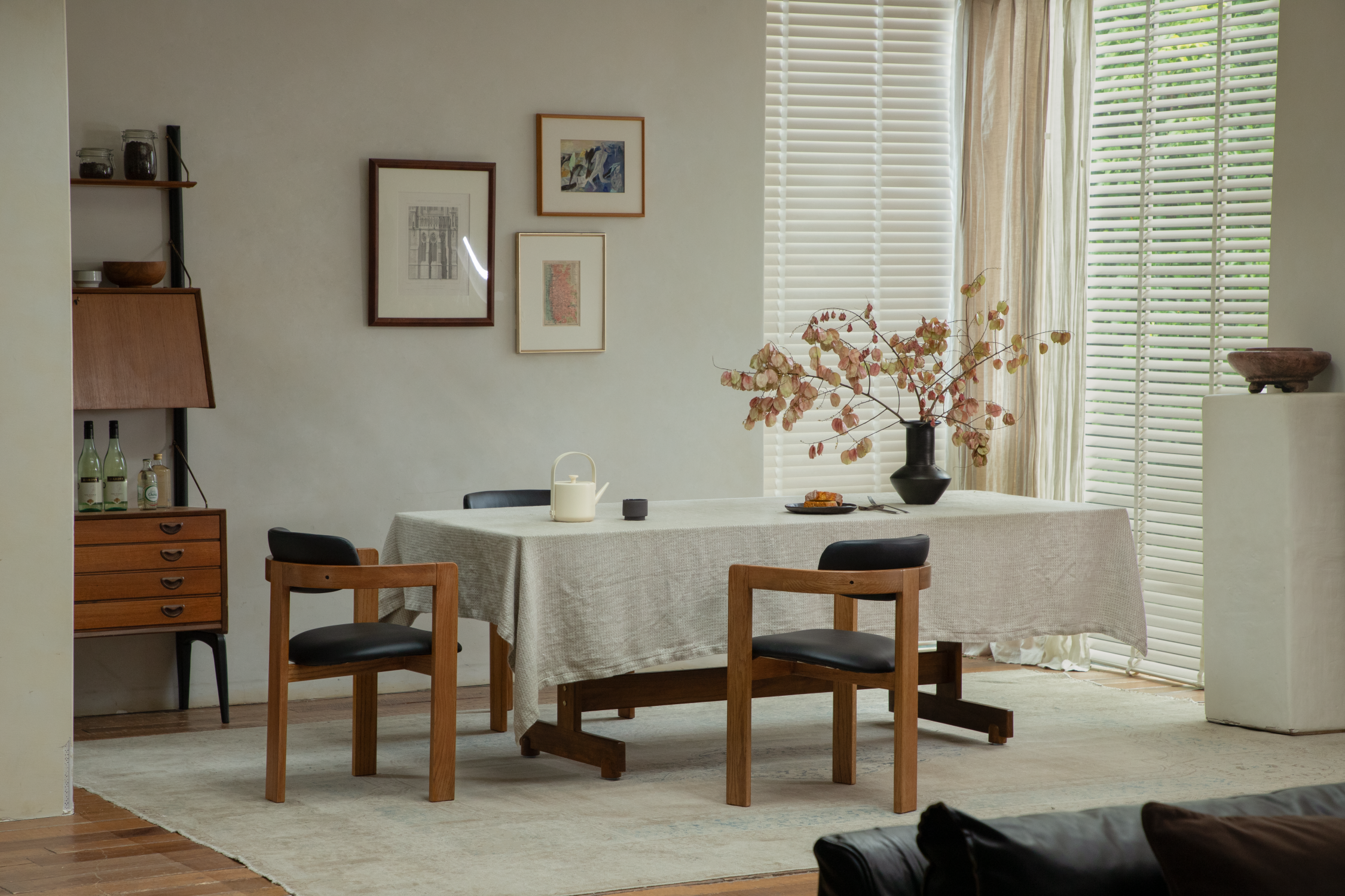 A minimalist dining room featuring a wooden table covered with a light cloth, modern wooden chairs with black seats, a wall-mounted shelving unit, and framed artwork in a serene, neutral-toned setting.