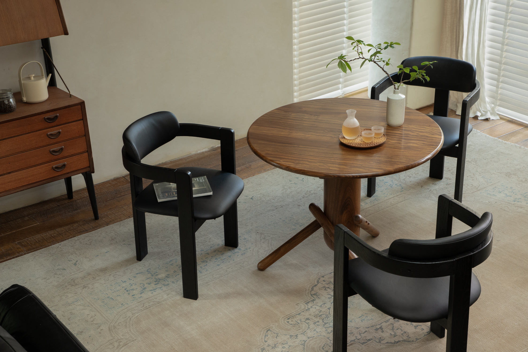 Round wooden dining table with black chairs, tableware, flowers, and soft daylight by the window
