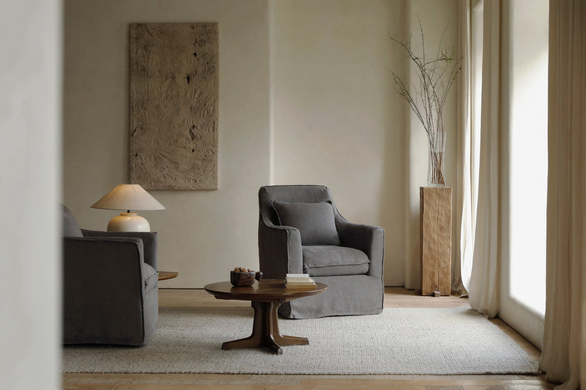 Two grey upholstered armchairs facing a round wooden coffee table on a light rug in a warm, minimalist room.