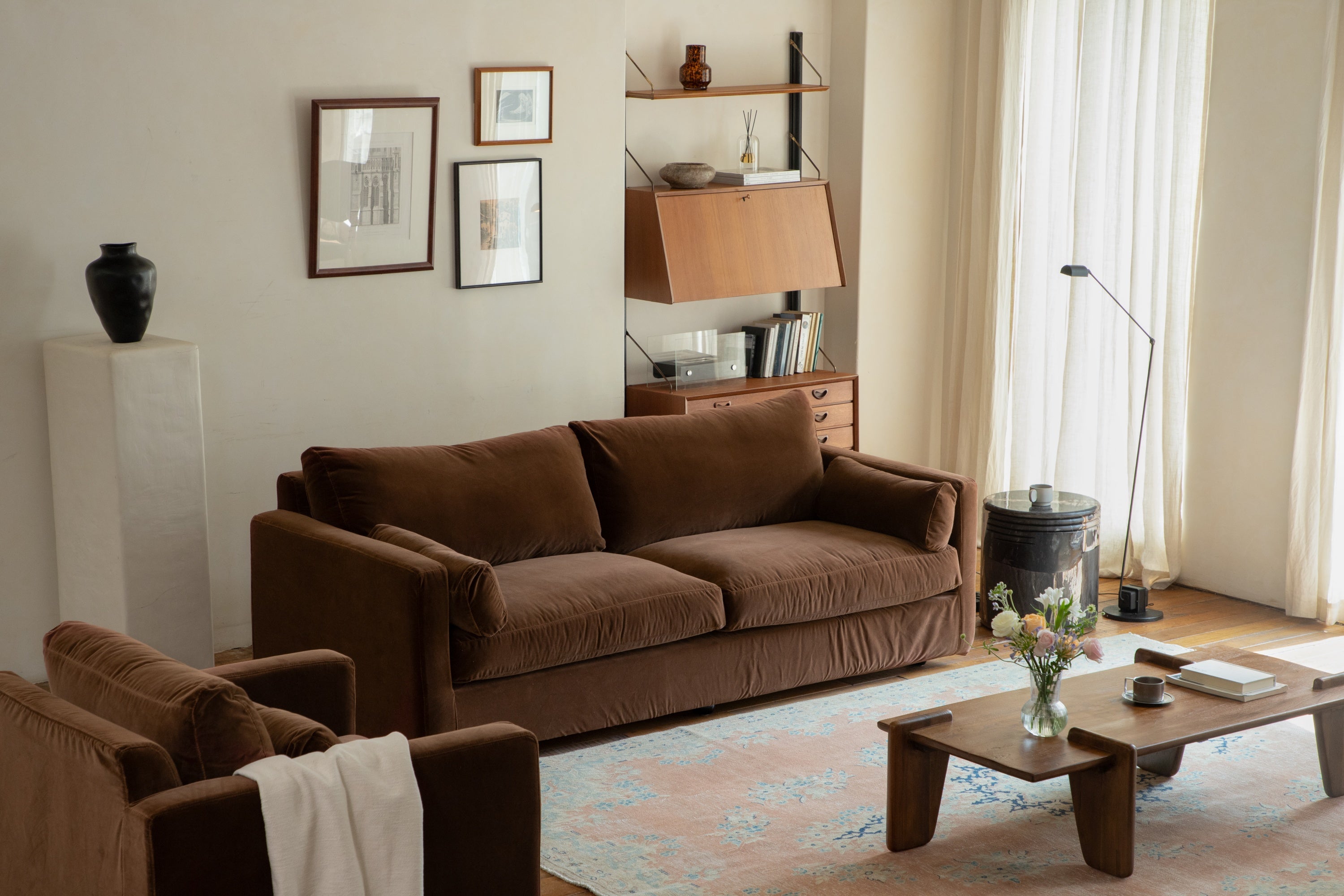 A brown sofa with a white blanket draped over one arm, positioned in a cozy living room with framed artwork on the wall and a bookshelf.
