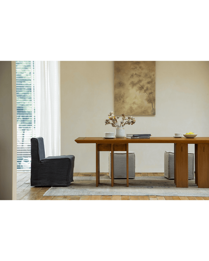 Teak dining table with decorative vase, fruit, and soft chairs in bright interior.