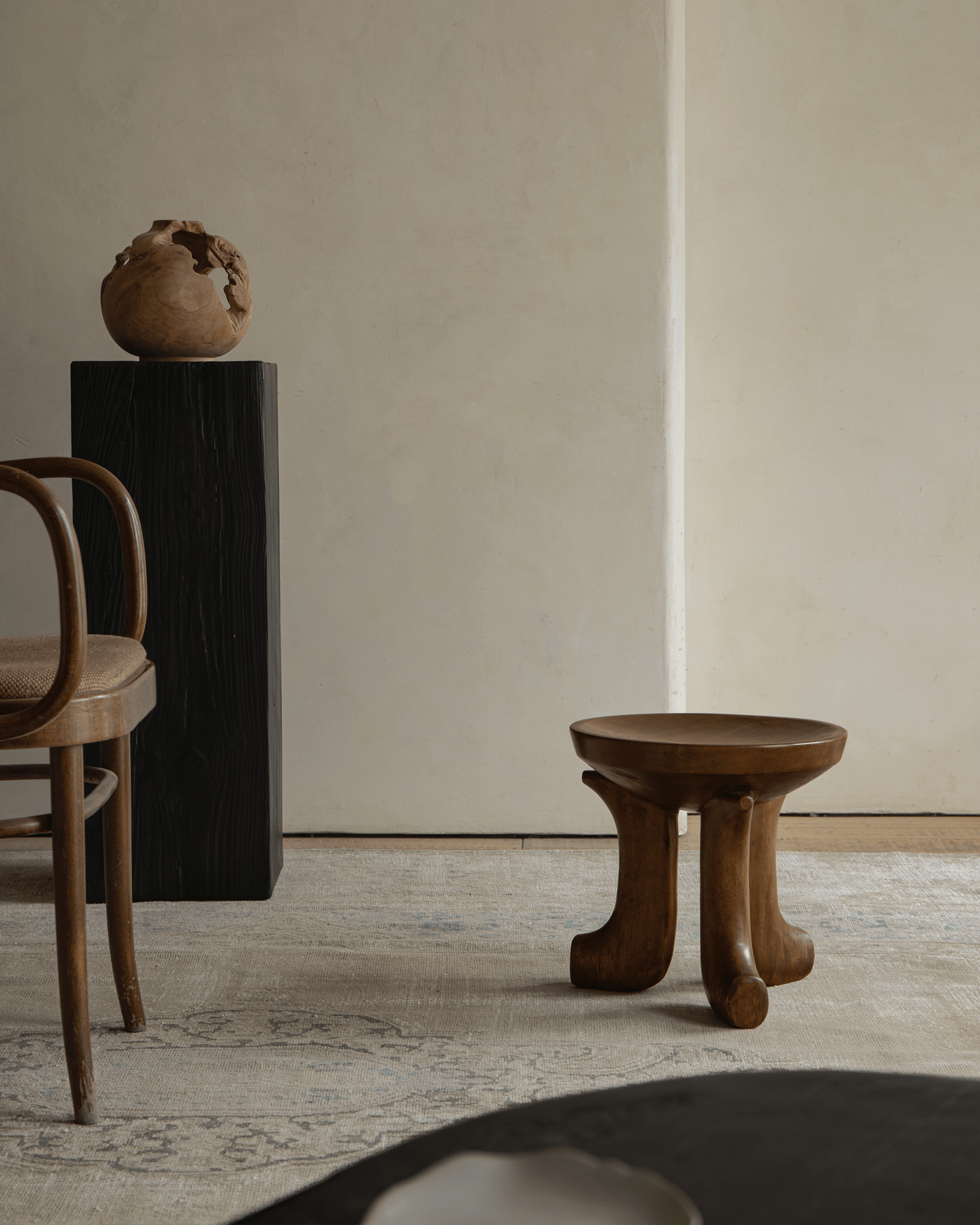 Quincy Stool with three legs, modern wooden design, natural finish. Featured in a cozy living room setting with wooden chair.