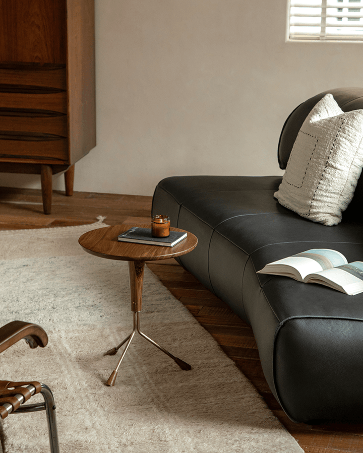 Larsson Side Table with round wood top and brass legs beside dark leather sofa