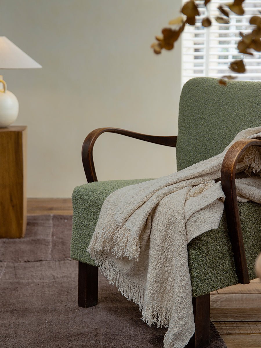 Cream cotton blanket on green armchair, showcasing texture and fringe, perfect for home decor.