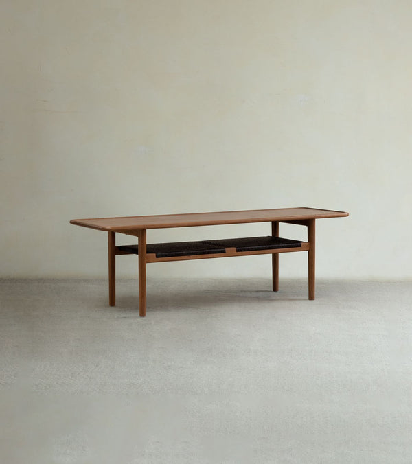 The Hans Accent Table by ZM Home is a rectangular wooden coffee table featuring tapered legs and a lower black woven shelf, displayed against a neutral wall and light floor.